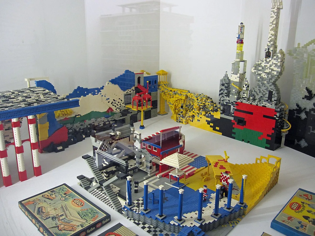 Een vitrine met grote bouwwerken van kleurige steentjes, waaronder bruggen, gebouwen en torens, met vooraan enkele oude LEGO System-dozen